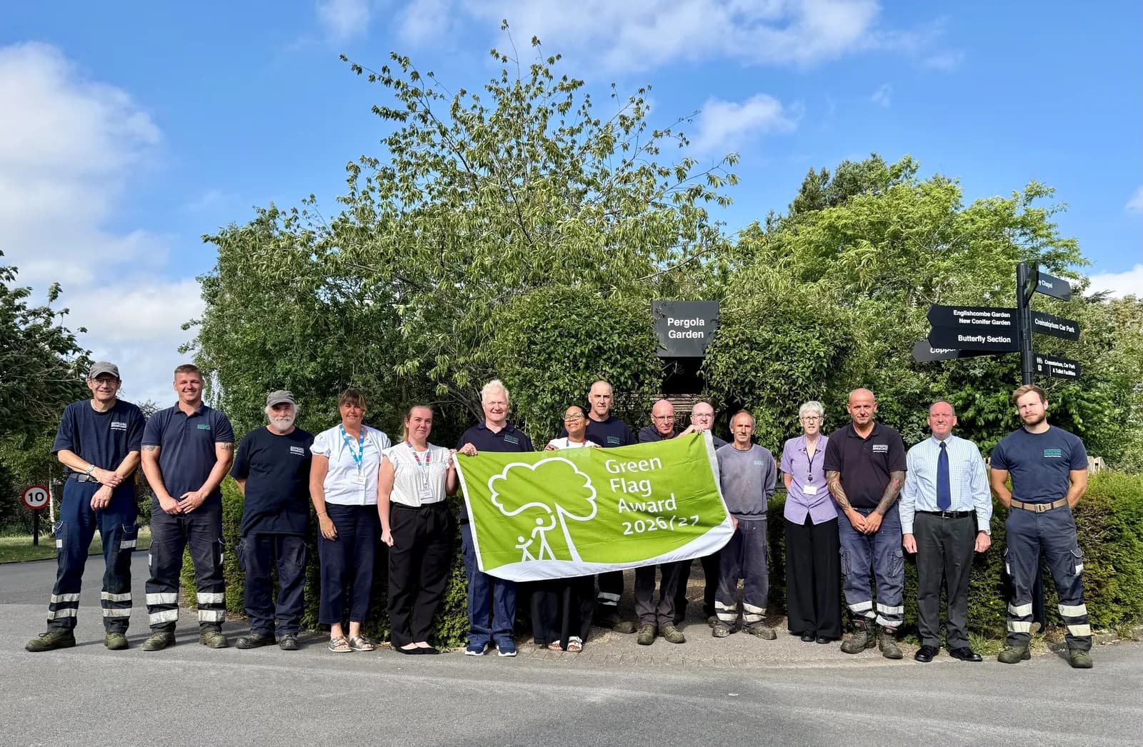 A diverse group of men and women stand smiling in front of a lush green background, holding a green banner that reads "Green Flag Award 2026/27."