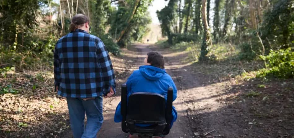 A man in a blue plaid shirt walks beside a person in a blue hoodie seated in a wheelchair on a dirt path through a wooded area.